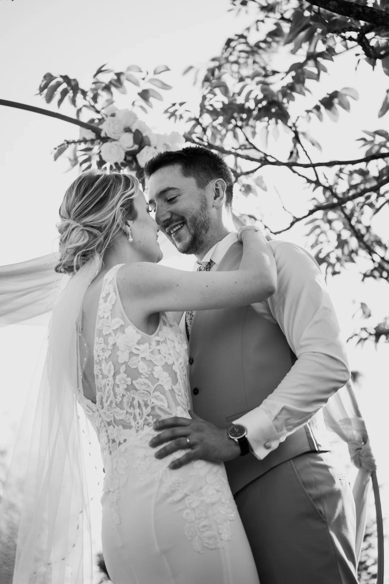 Les mariés sourient en se rapprochant sous les arbres, en noir et blanc